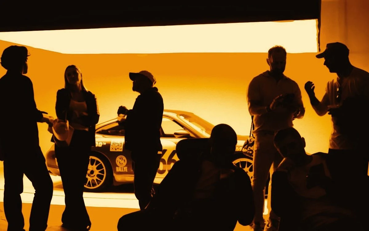 A group standing in silhouette in front of the P66 car under a light box, all in yellow light